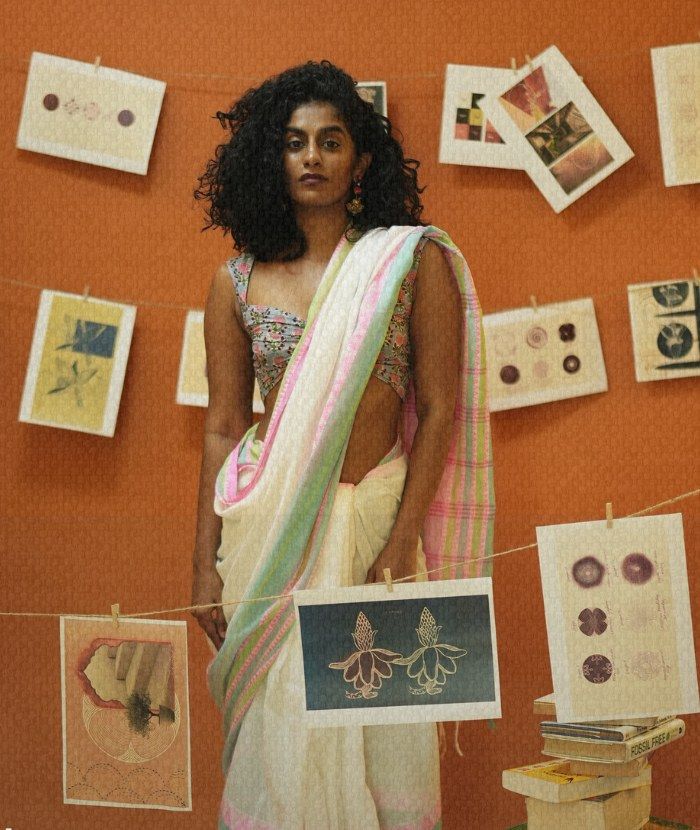 Placeholder portrait from the studio carousel set: a woman in a handloom sari standing in front of hand-drawn prints pegged on string against a terracotta wall.
