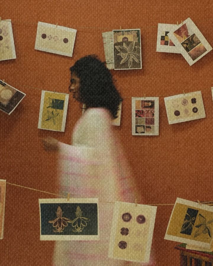 A woman in a white handloom sari moving past hand-drawn botanical prints pegged on string against a terracotta wall.